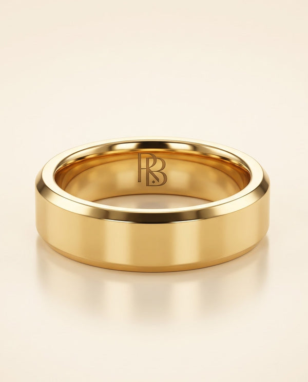 A polished yellow gold beveled tungsten band ring with a polished finish and an engraved 'RB' monogram inside, displayed on a warm cream background.