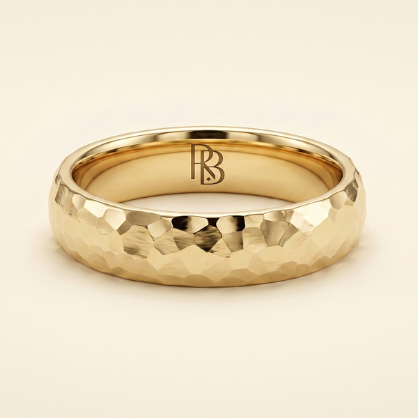 A gold band mens wedding ring with a hammered texture on the exterior and a polished interior by ring bear with comfort fit, set against a solid cream background.