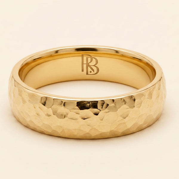 A yellow gold band mens wedding ring with a hammered texture on the exterior and a polished interior by ring bear with comfort fit, set against a solid cream background.