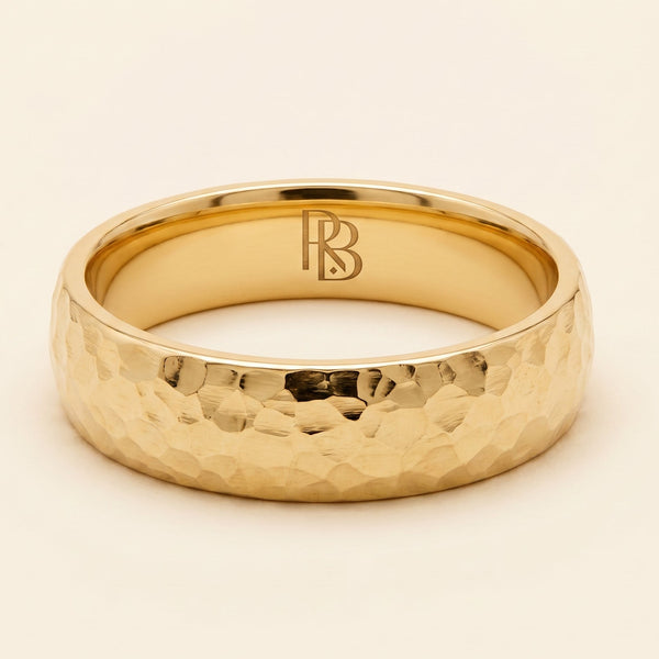 A yellow gold band mens wedding ring with a hammered texture on the exterior and a polished interior by ring bear with comfort fit, set against a solid cream background.