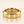 A polished yellow gold  beveled tungsten band ring with a polished finish and an engraved 'RB' monogram inside, displayed on a warm cream background.