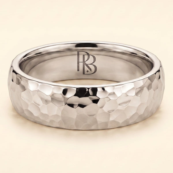 A white gold band mens wedding ring with a hammered texture on the exterior and a polished interior by ring bear with comfort fit, set against a solid cream background.