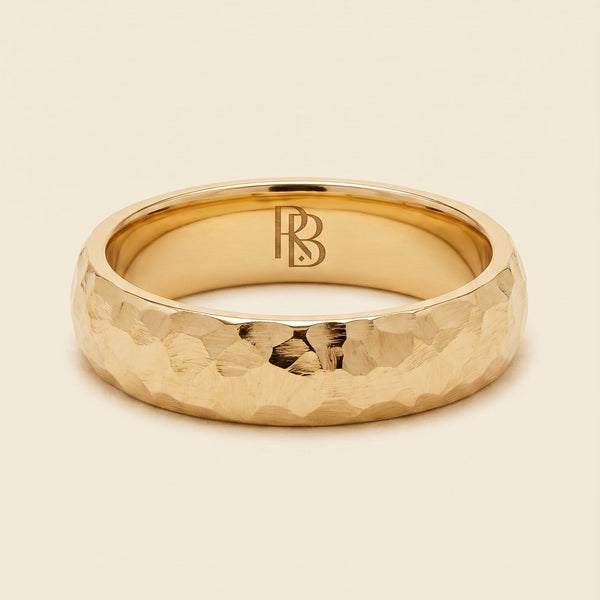 A yellow gold band mens wedding ring with a hammered texture on the exterior and a polished interior by ring bear with comfort fit, set against a solid cream background.
