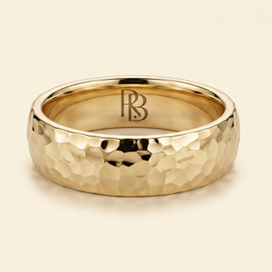 A gold band mens wedding ring with a hammered texture on the exterior and a polished interior by ring bear with comfort fit, set against a solid cream background.
