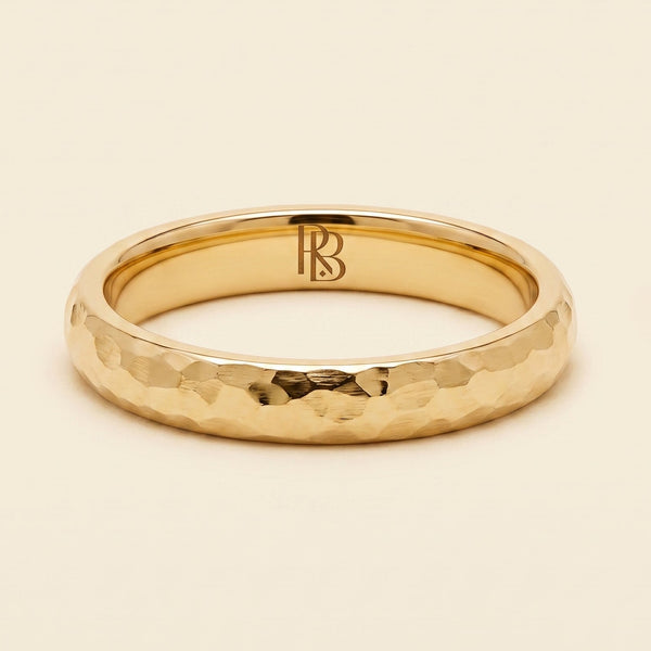 A yellow gold band mens wedding ring with a hammered texture on the exterior and a polished interior by ring bear with comfort fit, set against a solid cream background.
