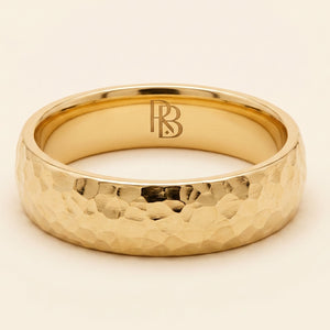 A yellow gold band mens wedding ring with a hammered texture on the exterior and a polished interior by ring bear with comfort fit, set against a solid cream background.